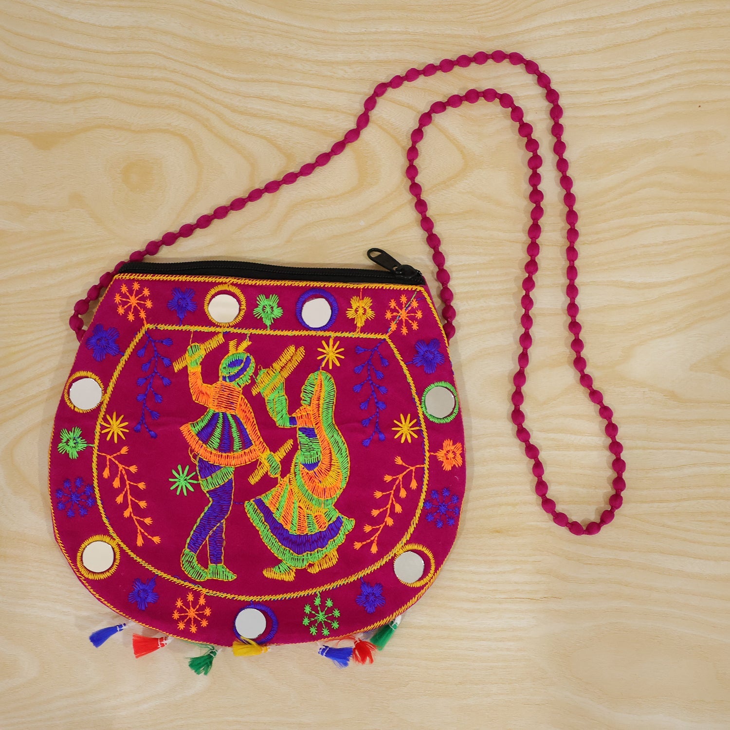 A vibrant pink sling bag with embroidered dancers, mirrors, and colorful tassels lies on a light wooden surface.