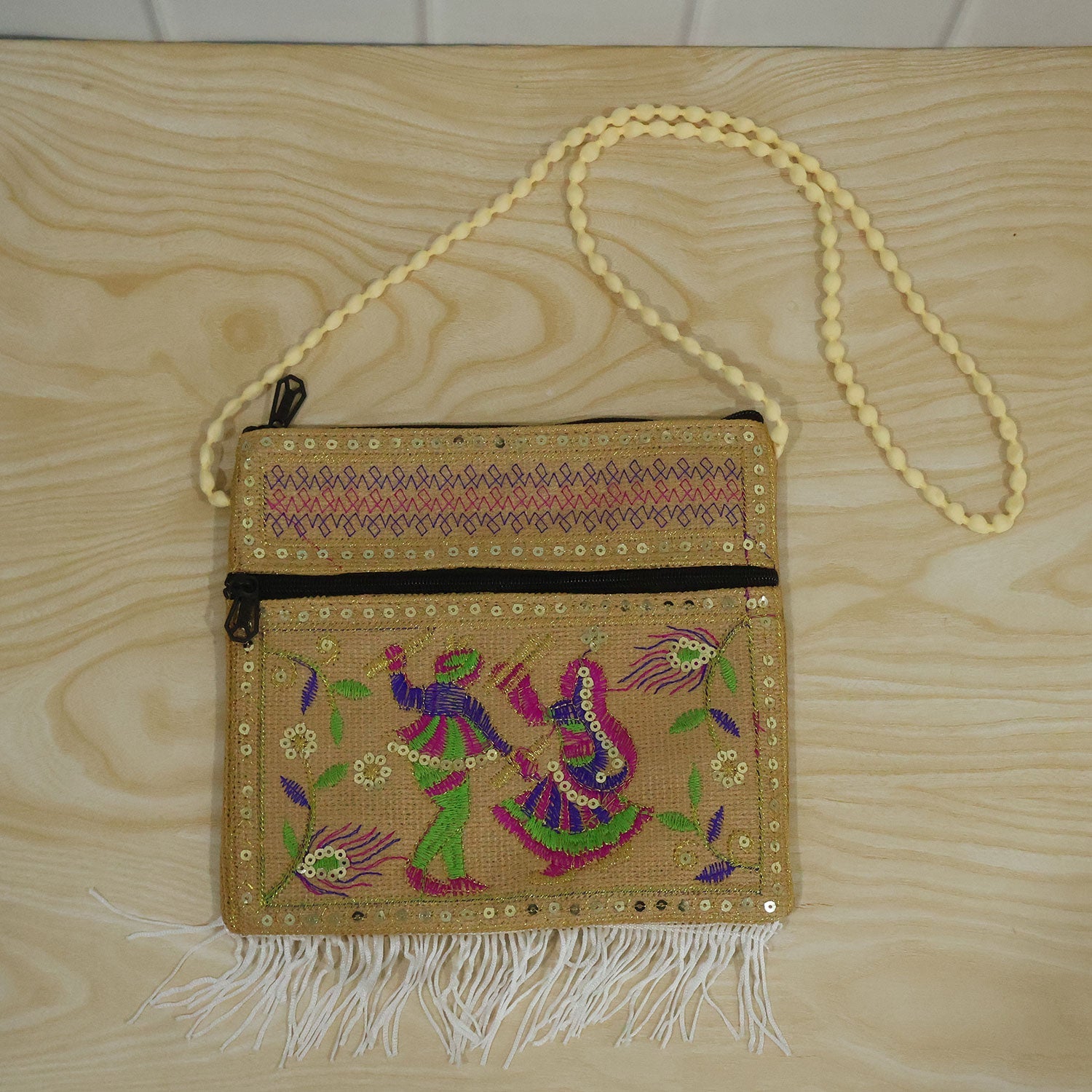 A beige jute sling bag with colorful embroidery, gold sequins, and white fringe on a wooden surface.