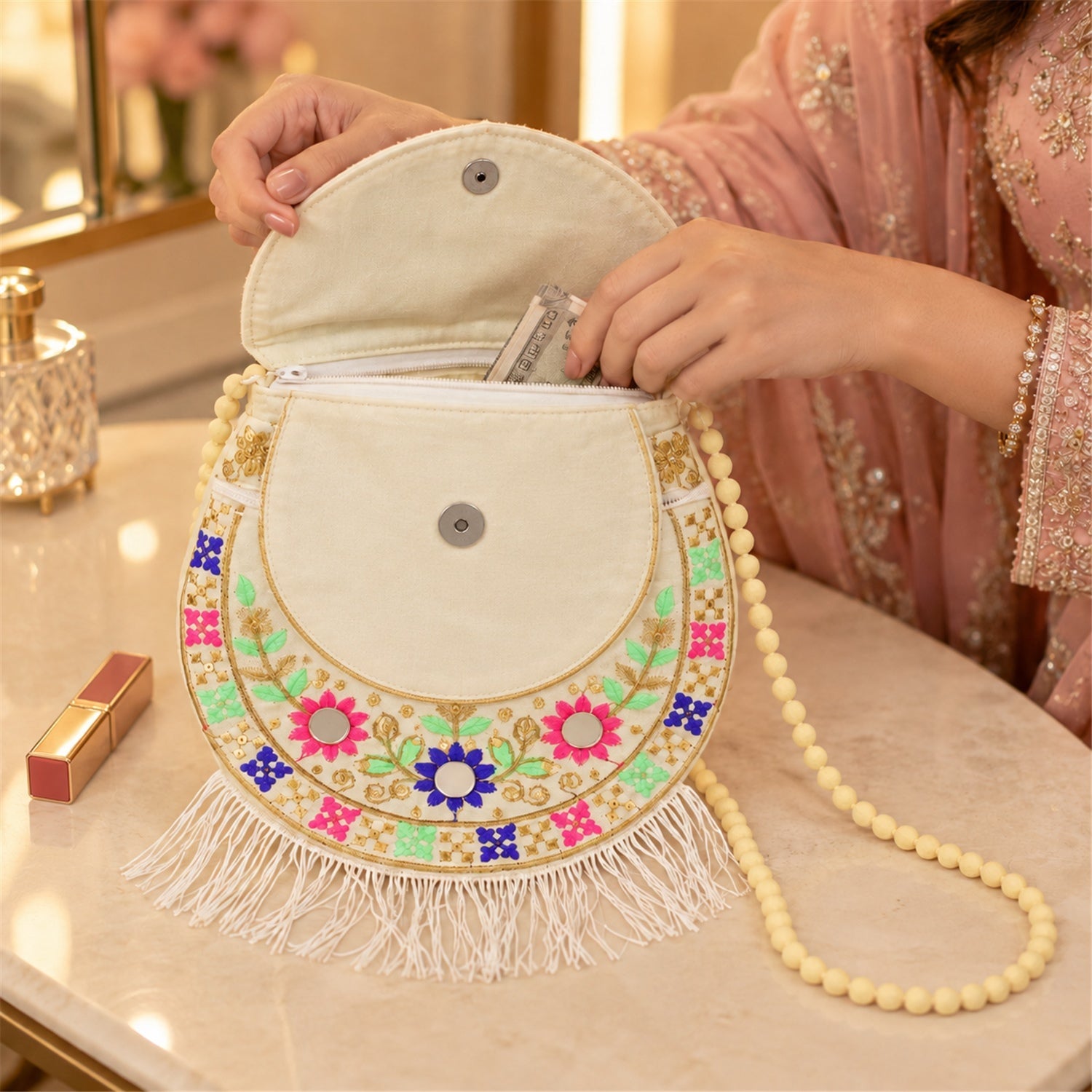 Hands open a white embroidered sling purse, revealing a zipper compartment with cash inside, on a vanity table.