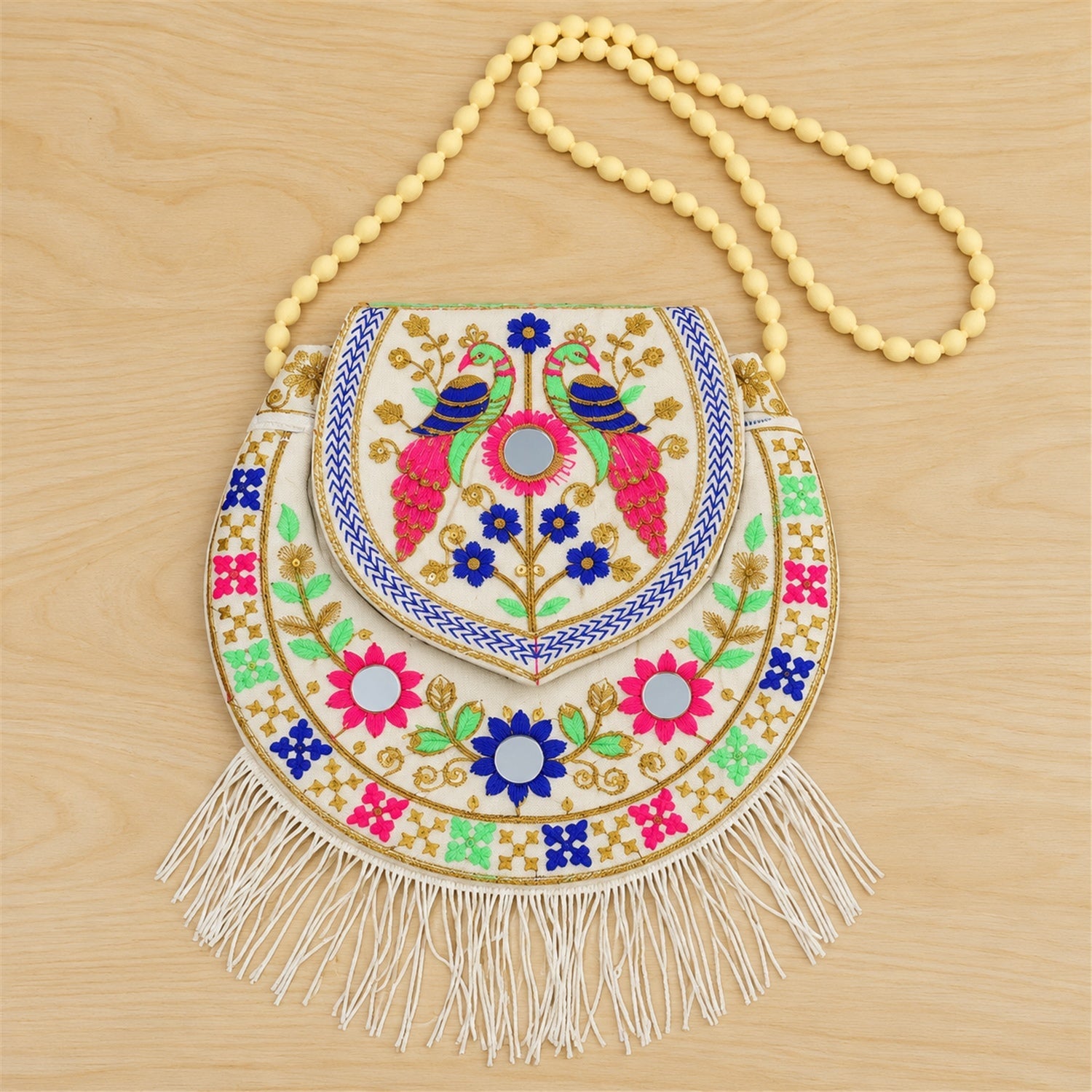 A white embroidered sling purse with colorful peacock and floral designs, mirror details, and fringe on a wooden surface.
