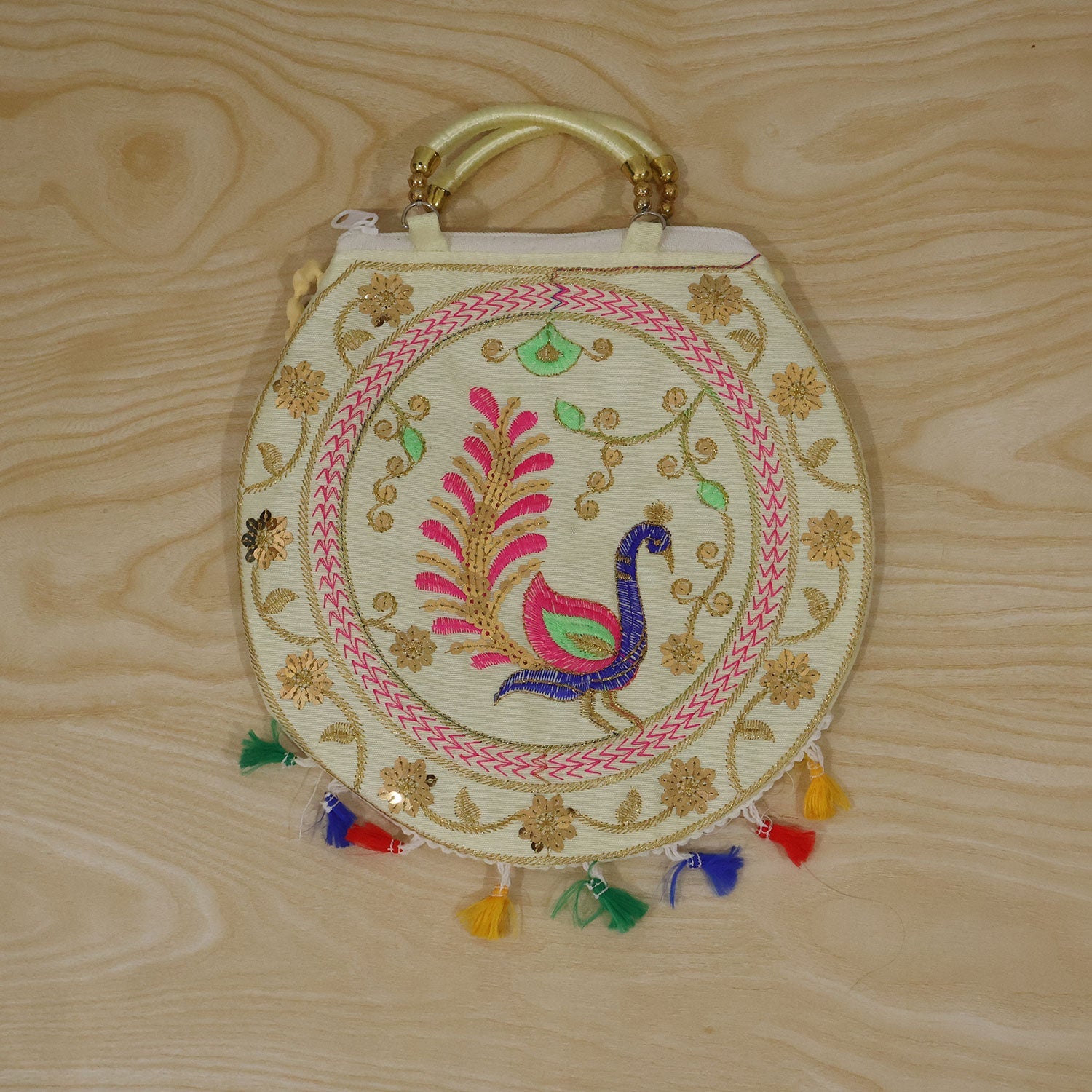 Overhead view of a cream round bag with a colorful embroidered peacock design and multi-colored tassels on a wooden surface.