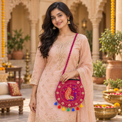 A woman in a peach outfit smiles while holding a round fuchsia purse with colorful tassels and peacock embroidery.