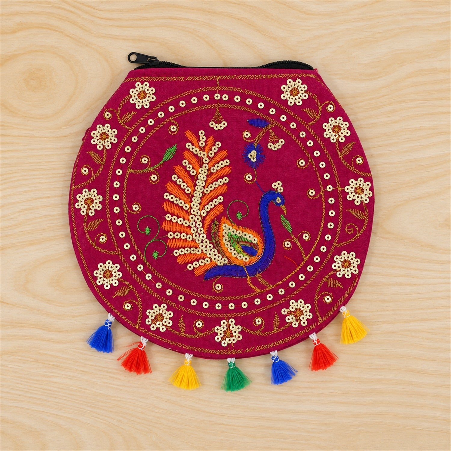 A fuchsia round purse with peacock embroidery, sequins, and colorful tassels, lying flat on a light wooden surface.