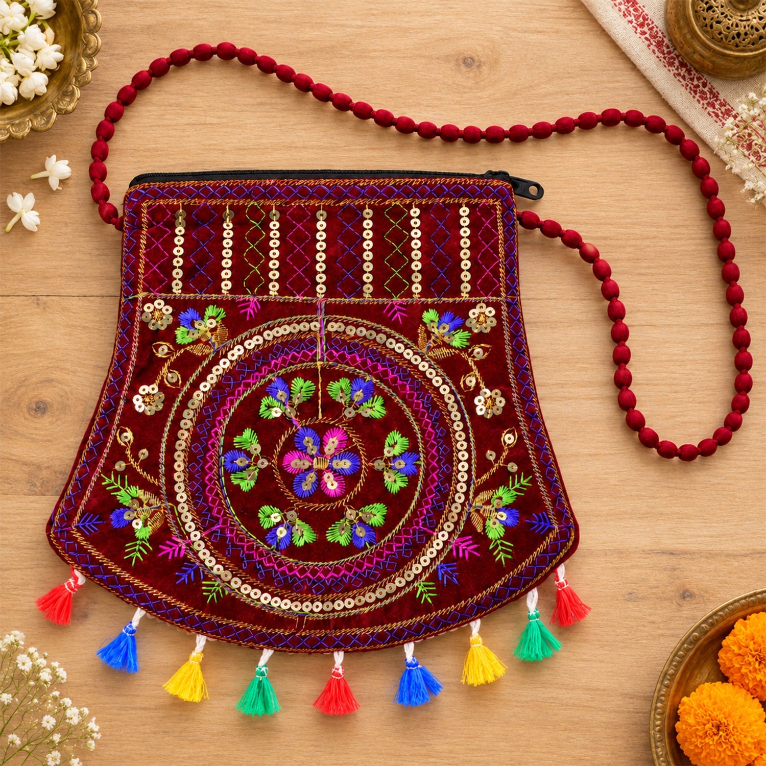 A maroon velvet sling bag with intricate colorful embroidery, sequins, and vibrant tassels lies on a wooden surface.