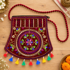 A maroon velvet sling bag with intricate colorful embroidery, sequins, and vibrant tassels lies on a wooden surface.