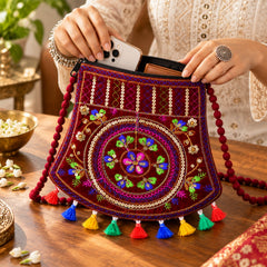 A person places a phone into a maroon velvet sling bag with colorful embroidery and tassels, showing its capacity.