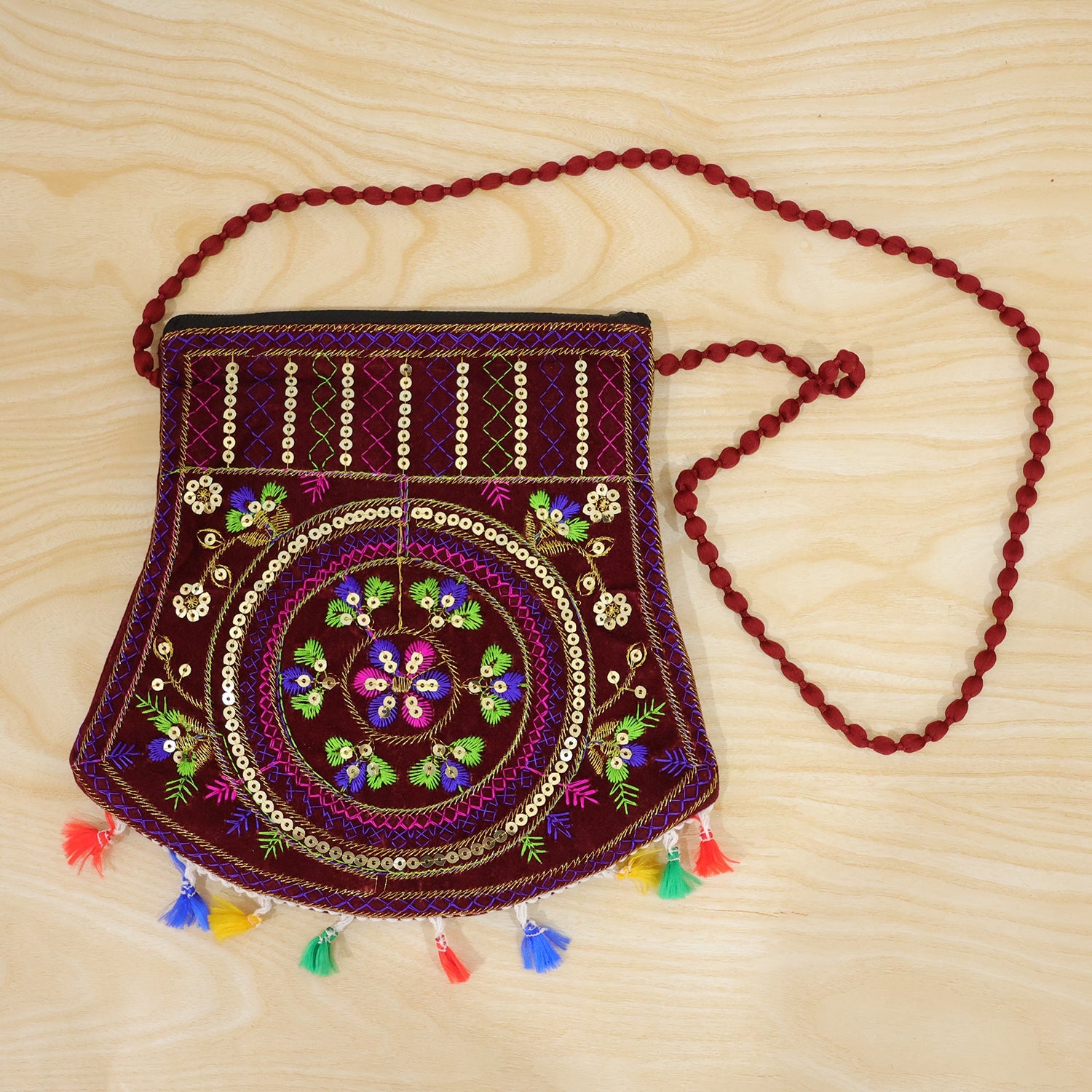 A maroon velvet sling bag with a long red beaded strap, colorful embroidery, and tassels on a light wooden surface.