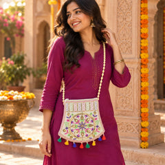 A smiling woman in a magenta dress wears an embroidered jute sling purse with colorful tassels and a beaded strap.