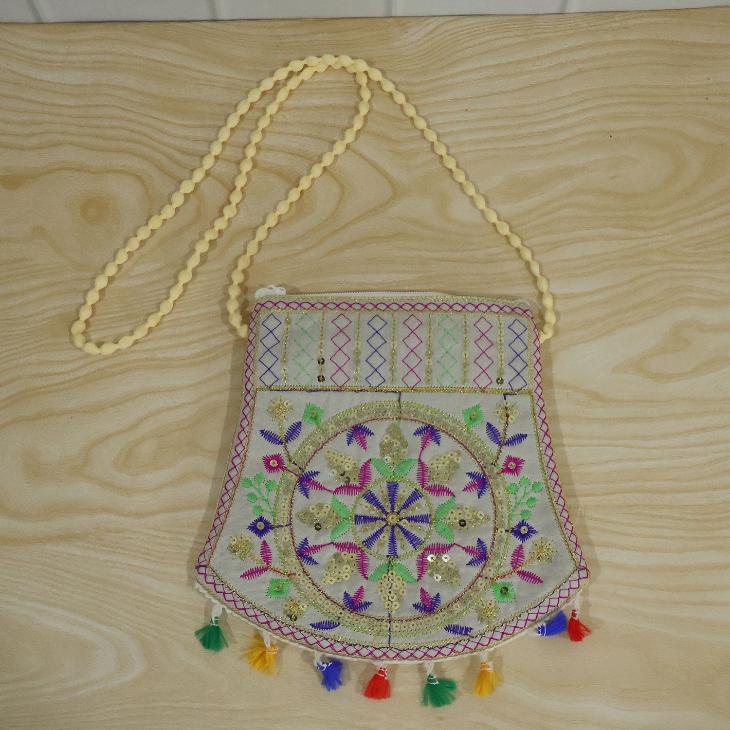 The embroidered jute sling purse with a beaded strap and colorful tassels lies flat on a light wood surface.