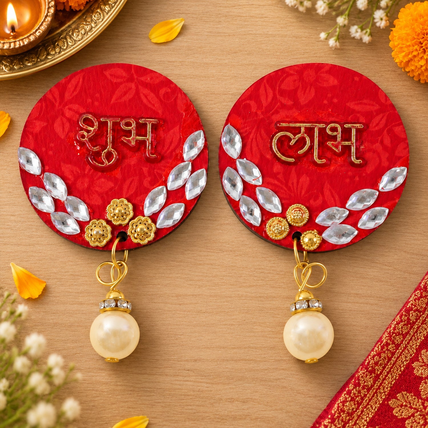A pair of red circular Shubh Labh decor pieces with gold details and pearl drops rest on a light wooden surface next to a