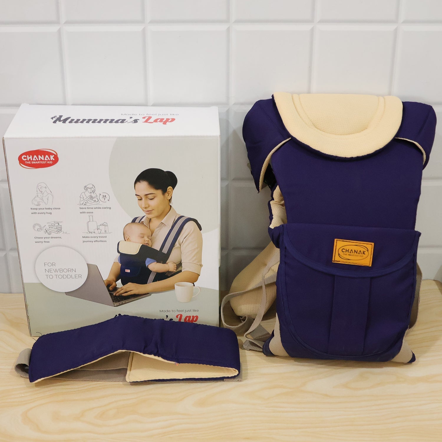 A navy and beige baby carrier, its box, and a detached waist belt are arranged on a light wooden surface.