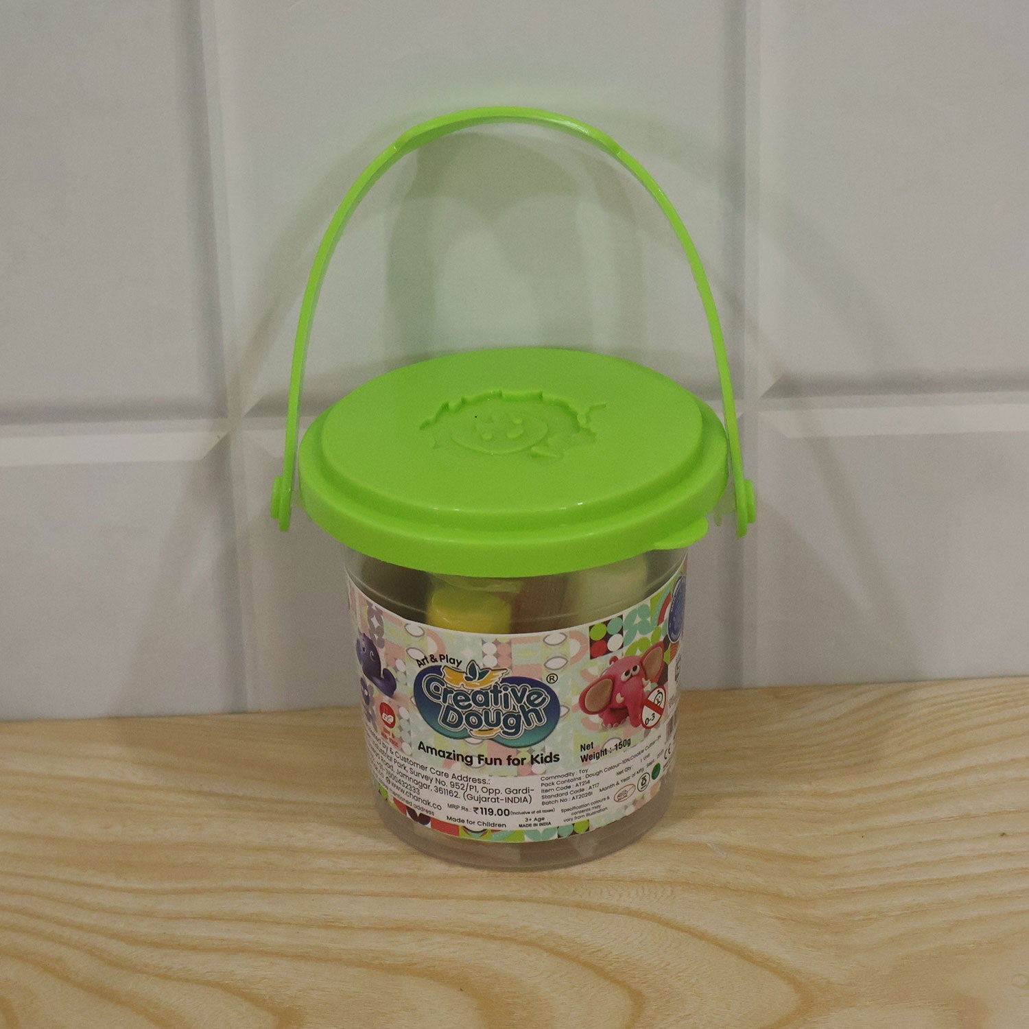 A clear plastic bucket with a green lid and handle, filled with colorful dough, sits on a light wooden surface.