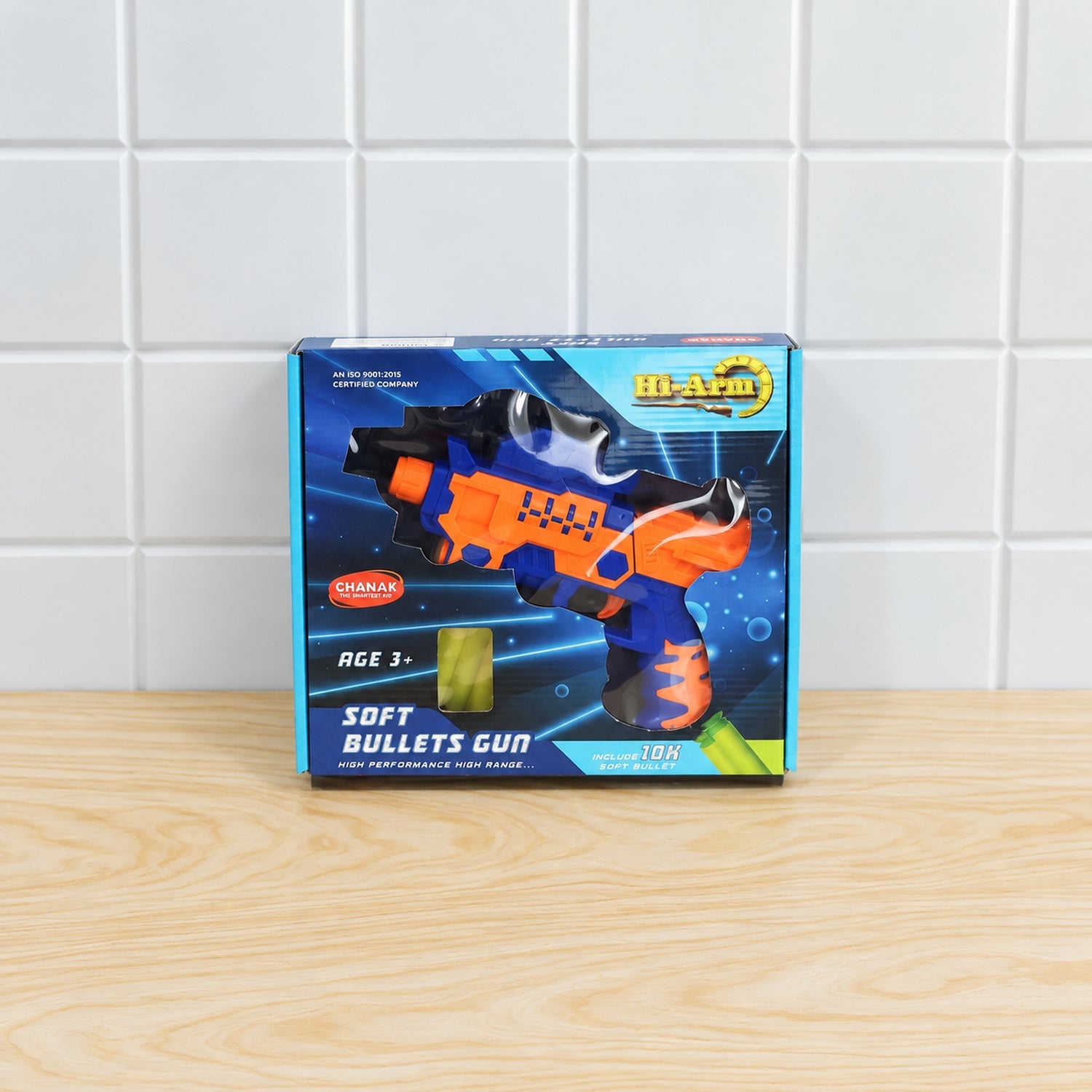 The product box for an orange and blue soft bullets toy gun is centered on a light wooden surface against a white tiled wall.