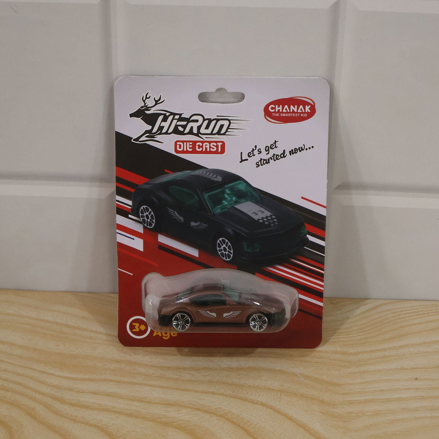 A single brown die-cast toy car in its blister packaging, hanging against a white wooden wall.