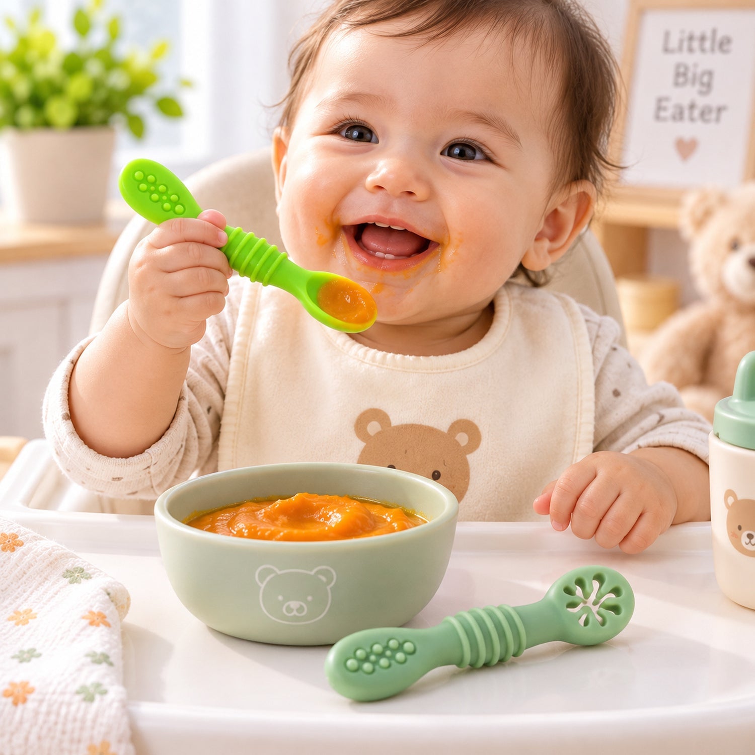 A smiling baby in a high chair holding a green silicone spoon, with a bowl of pureed food and another spoon on the tray.