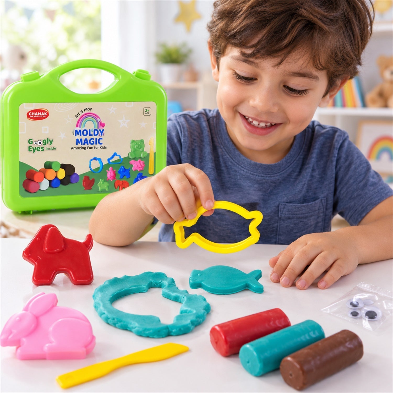 A boy smiles while holding a brown clay fish, with a green case, colorful clay, and plastic moulds on the table.
