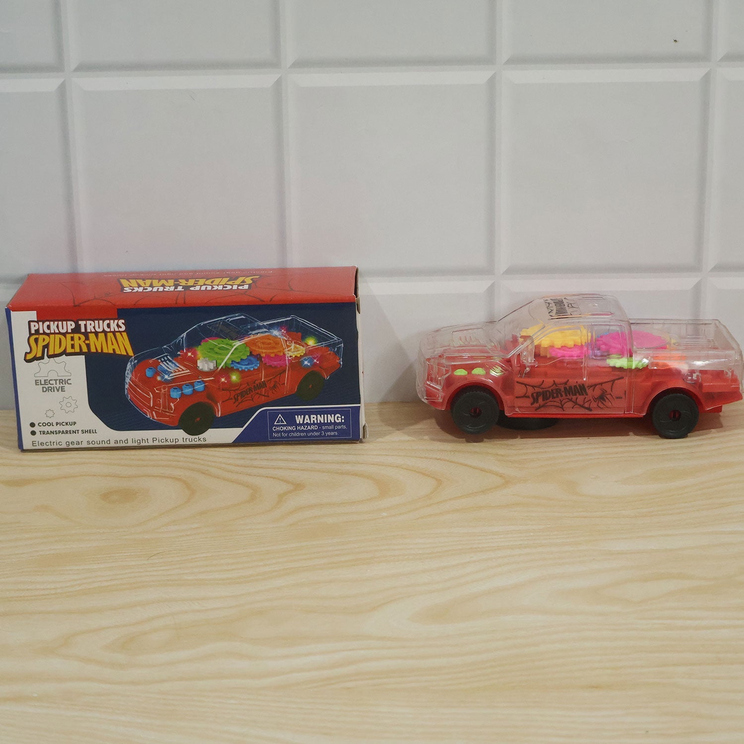 A transparent red toy pickup truck sits next to its red and blue packaging box on a light wood surface.