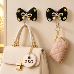 Two black bow-shaped hooks with gold accents and a gold bow, holding a white handbag and a pink quilted pouch.
