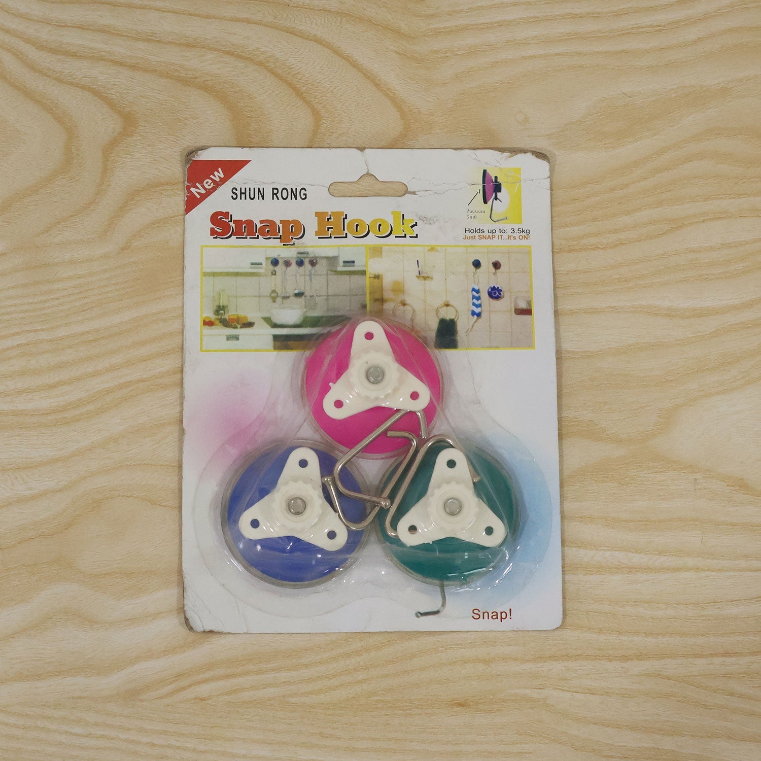 Three colorful suction cup snap hooks in their original retail packaging on a light wooden surface.