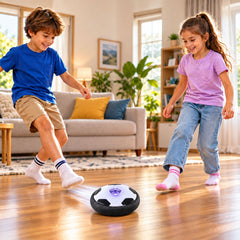 Two children play with a white and black hoverball soccer toy on a wooden floor in a sunlit living room.