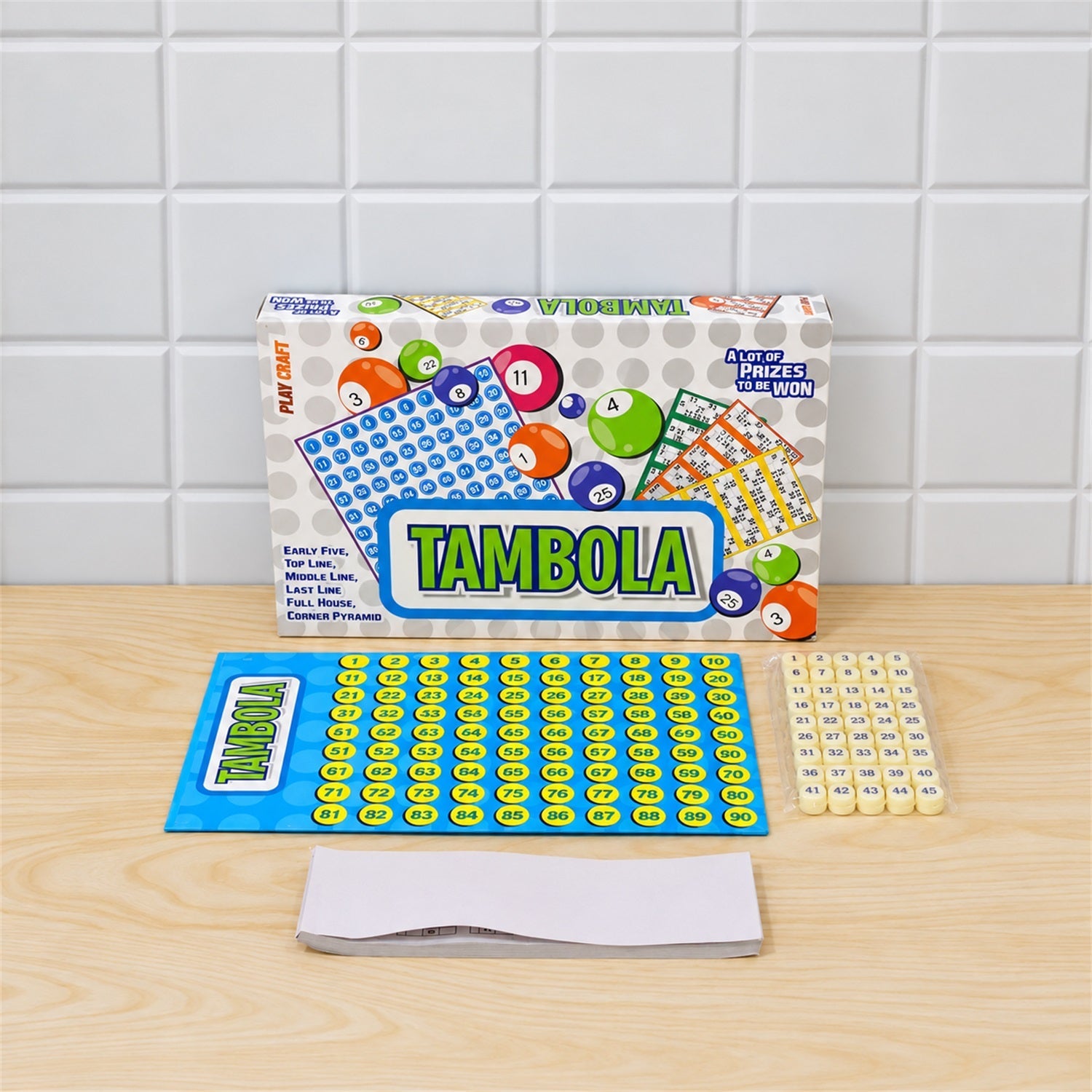 The Tambola game set is arranged on a light wooden surface against a white tiled wall, showing the box, board, tokens, and