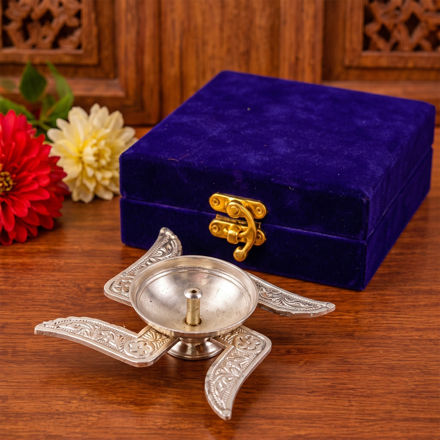 A silver swastik diya with intricate patterns sits on a wooden surface, next to a closed blue velvet box.