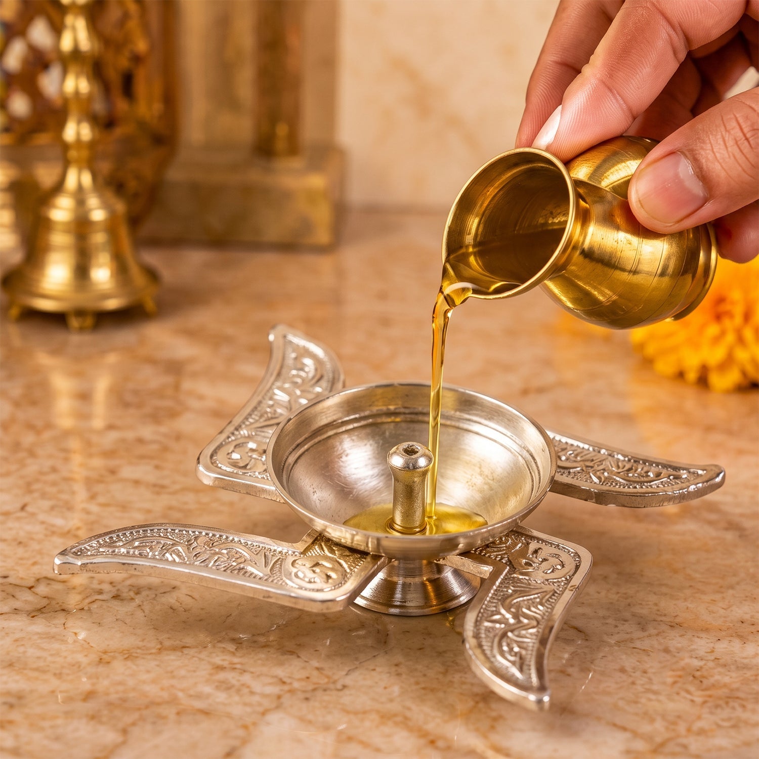 A hand pours golden oil from a small brass pot into the silver swastik diya on a light brown marble surface.