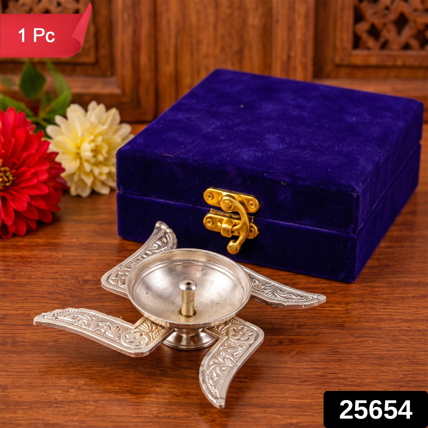 A silver swastik diya with detailed engravings rests on wood, beside a blue velvet box and red flowers.