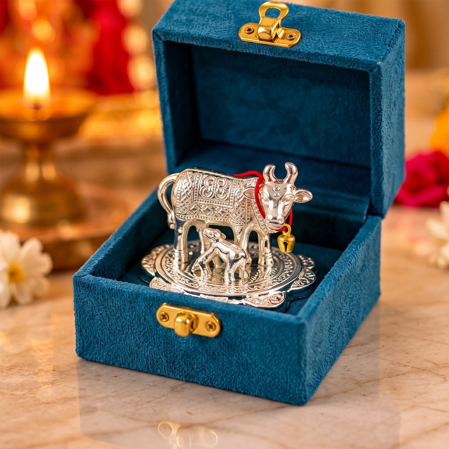 A silver Kamdhenu cow and calf idol with a red string and bell, inside an open teal velvet box.