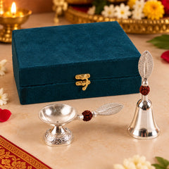 A silver finish pooja bell and diya set with leaf handles and rudraksha beads, displayed next to a teal velvet box.