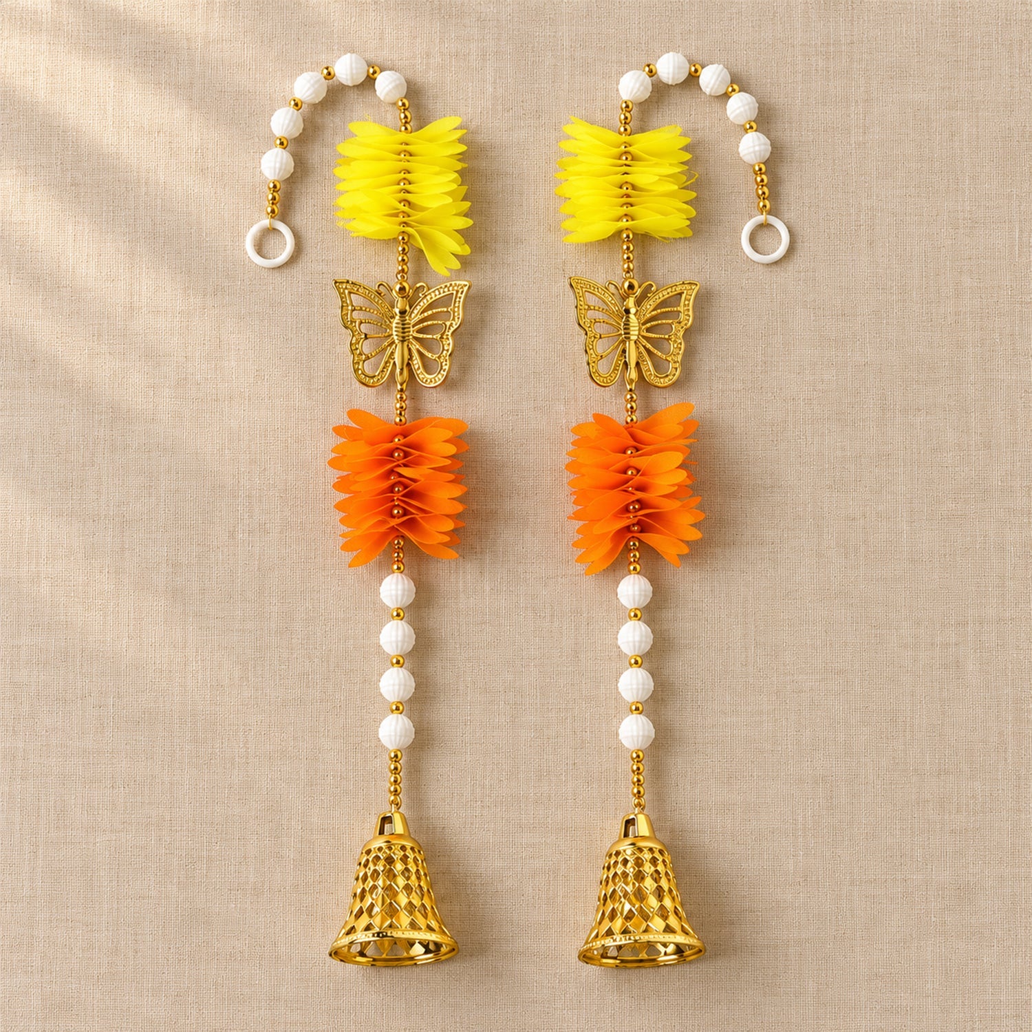 Two ornate door hangings, each with yellow and orange paper flowers, a golden butterfly, and a bell, laid flat on a textured
