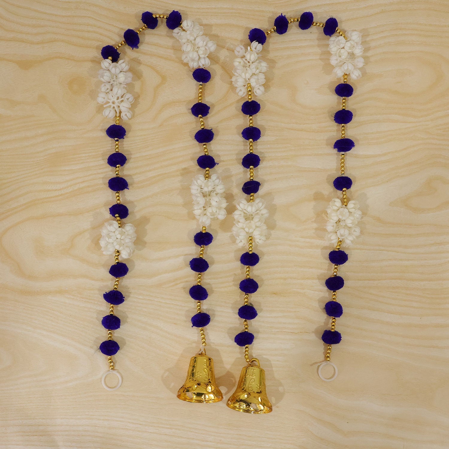 Four decorative strands, each with blue pom-poms, white floral clusters, and a golden bell, are arranged on a light wooden