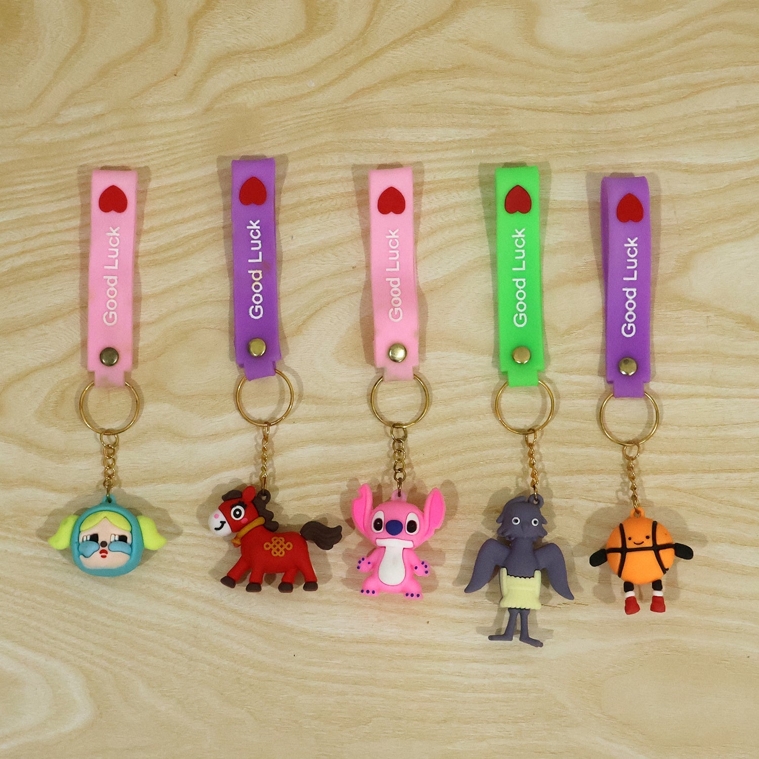 Five unique cartoon character keychains with colorful silicone straps and gold rings are laid out on a light brown wooden