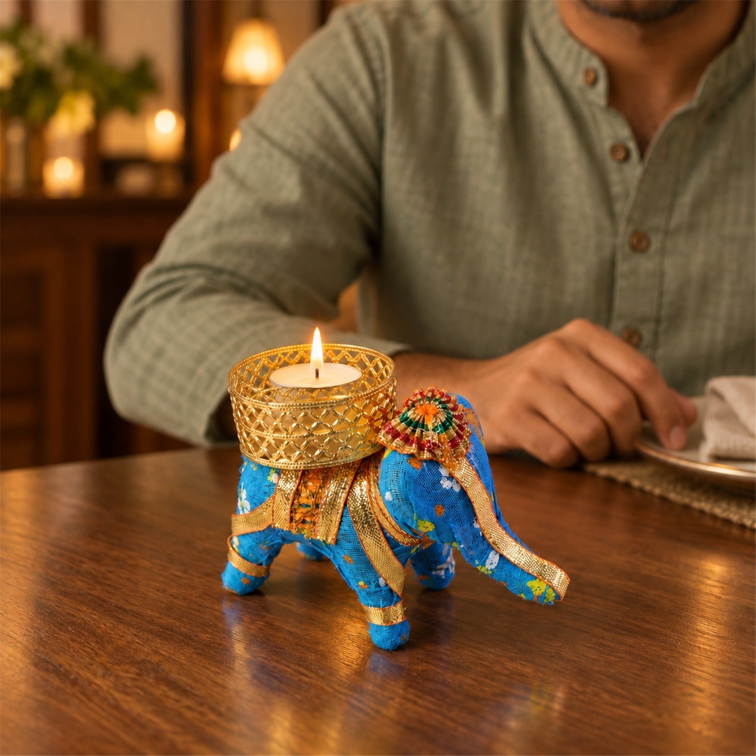 A blue fabric elephant candle holder with a lit tealight on a wooden table, a person seated in the background.