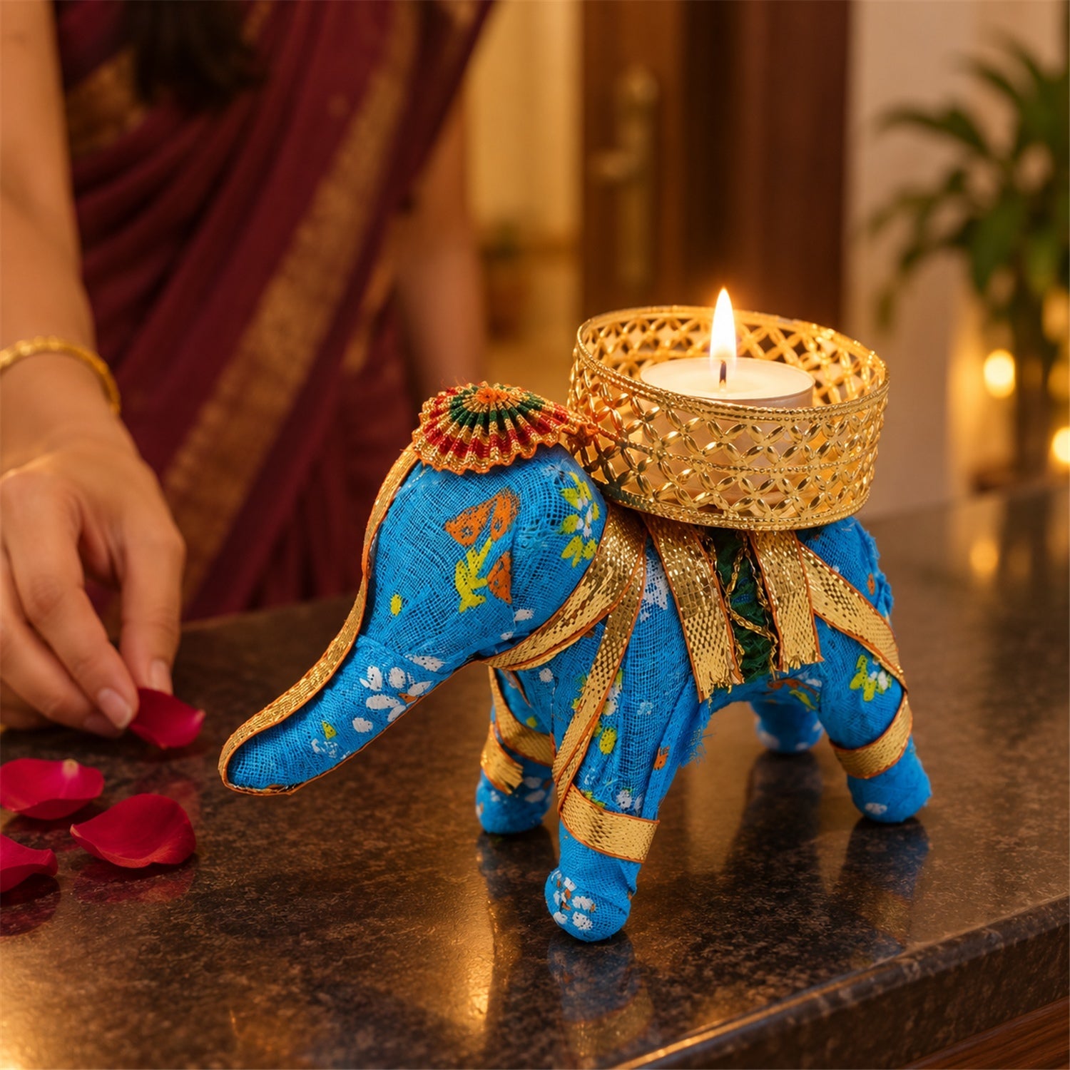A blue fabric elephant candle holder with a lit tealight, a person's hands placing red petals on a dark surface.