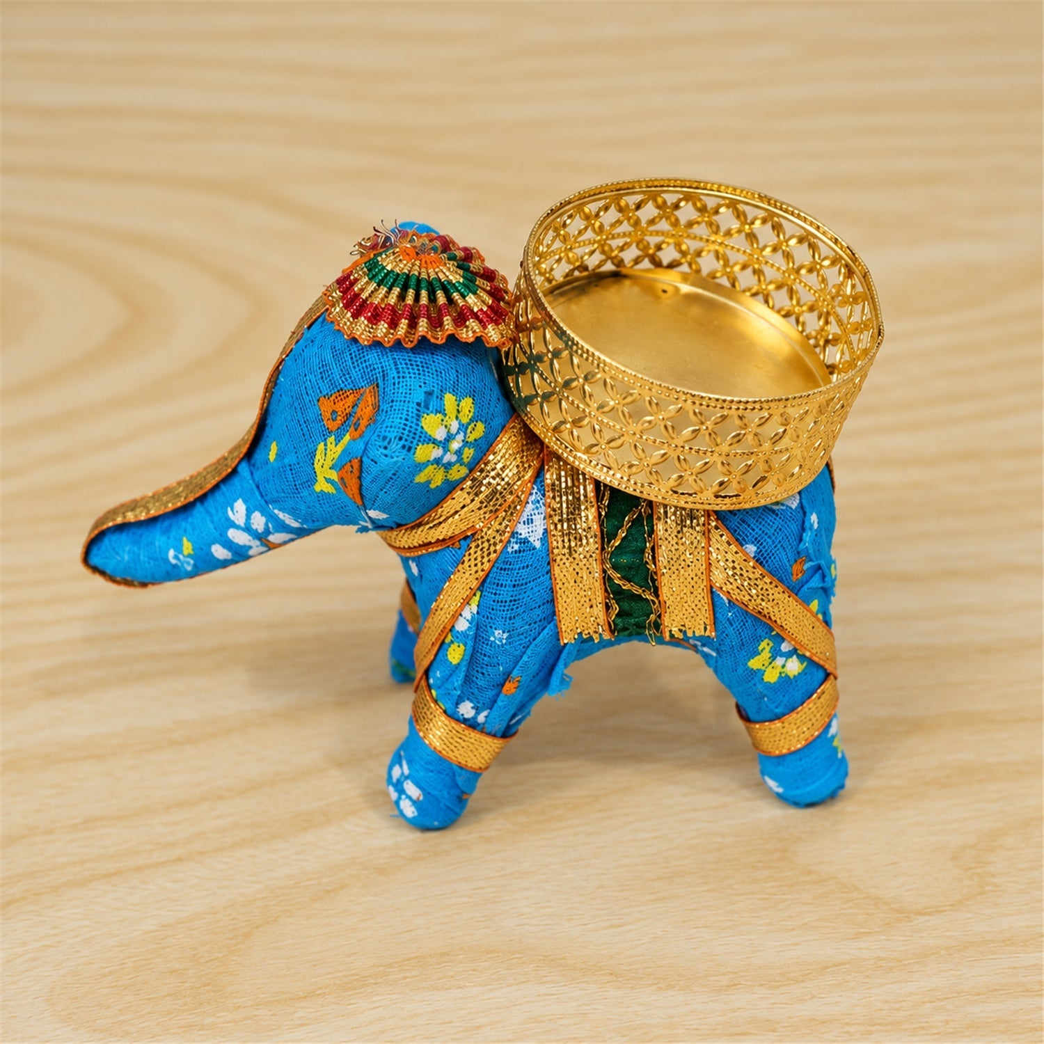 A blue fabric elephant candle holder with an empty gold lattice cup, positioned on a light wooden surface.