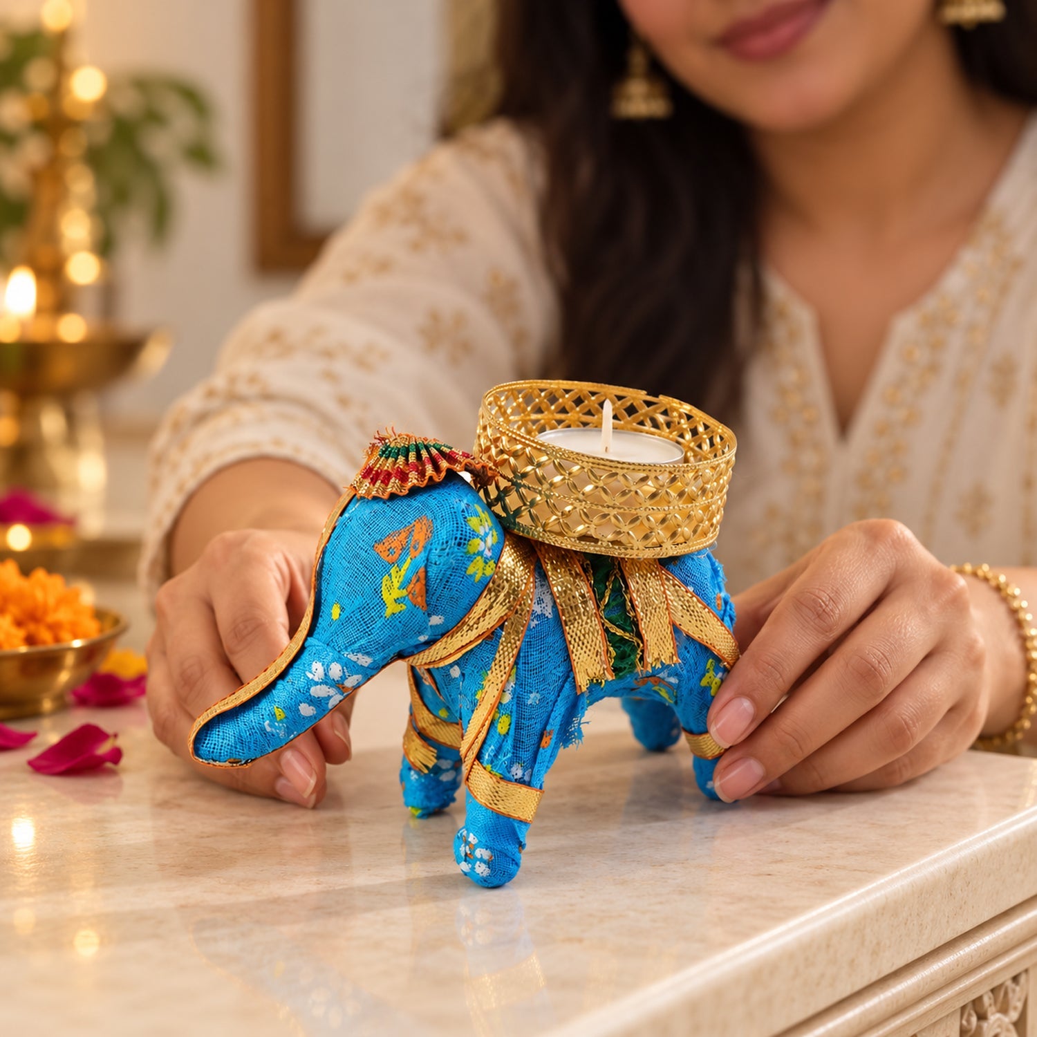 A blue fabric elephant candle holder with a lit tealight, a person's hands gently touching the table near it.