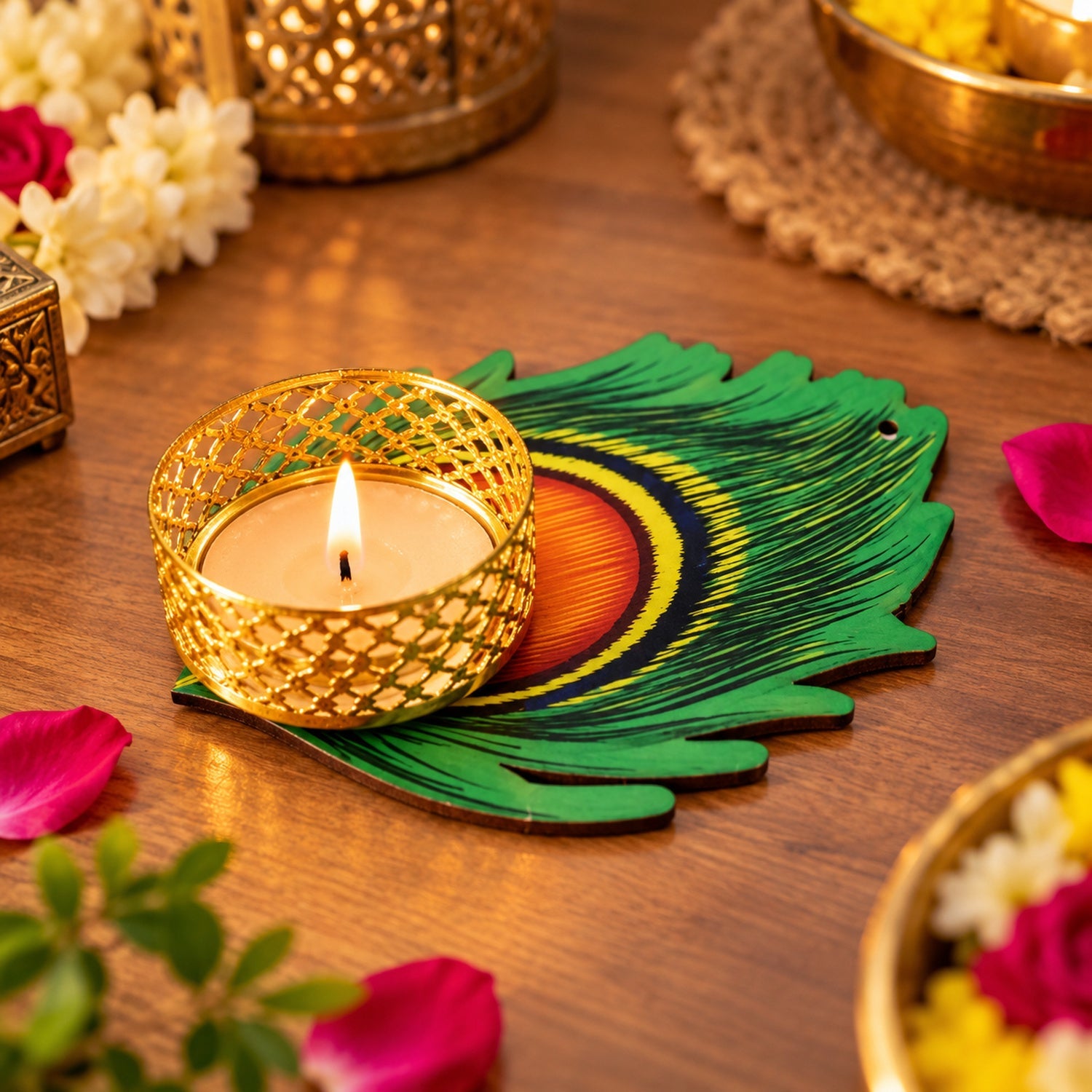 A close-up of a lit tealight in a gold filigree holder on a green peacock feather base, with blurred flowers.
