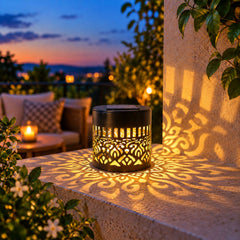 A black solar lamp with a cut-out mandala pattern casts a warm glow on a patio ledge at dusk.