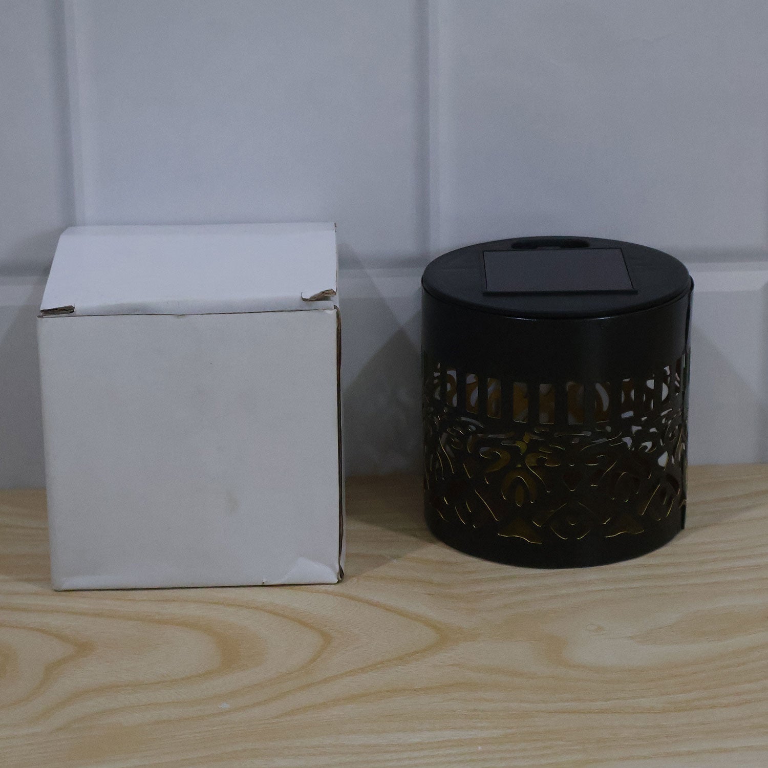 A black solar lamp with a cut-out mandala pattern next to a white box on a wooden surface.
