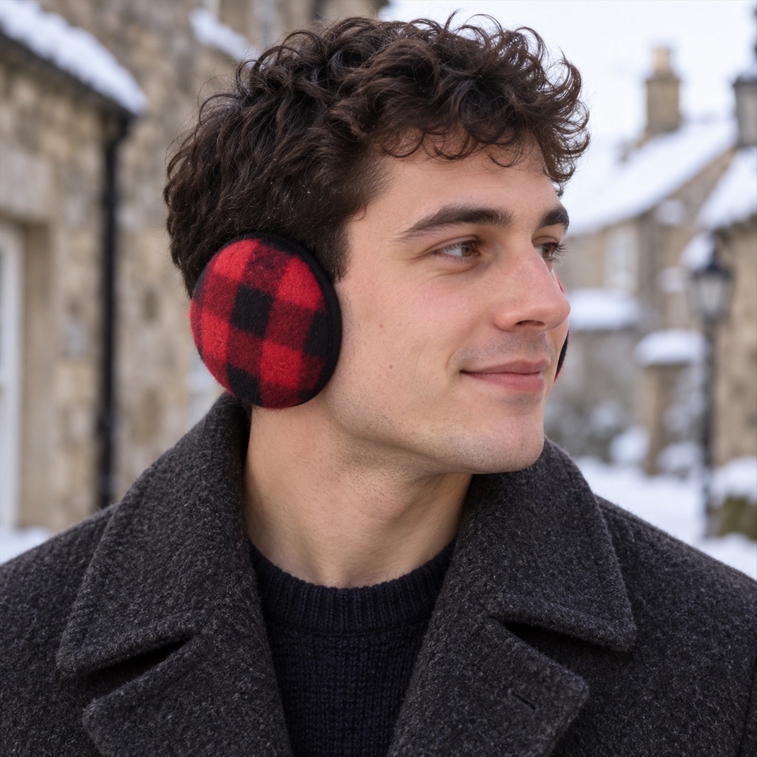 A man with curly hair wears red and black plaid ear warmers and a gray coat in a snowy village.