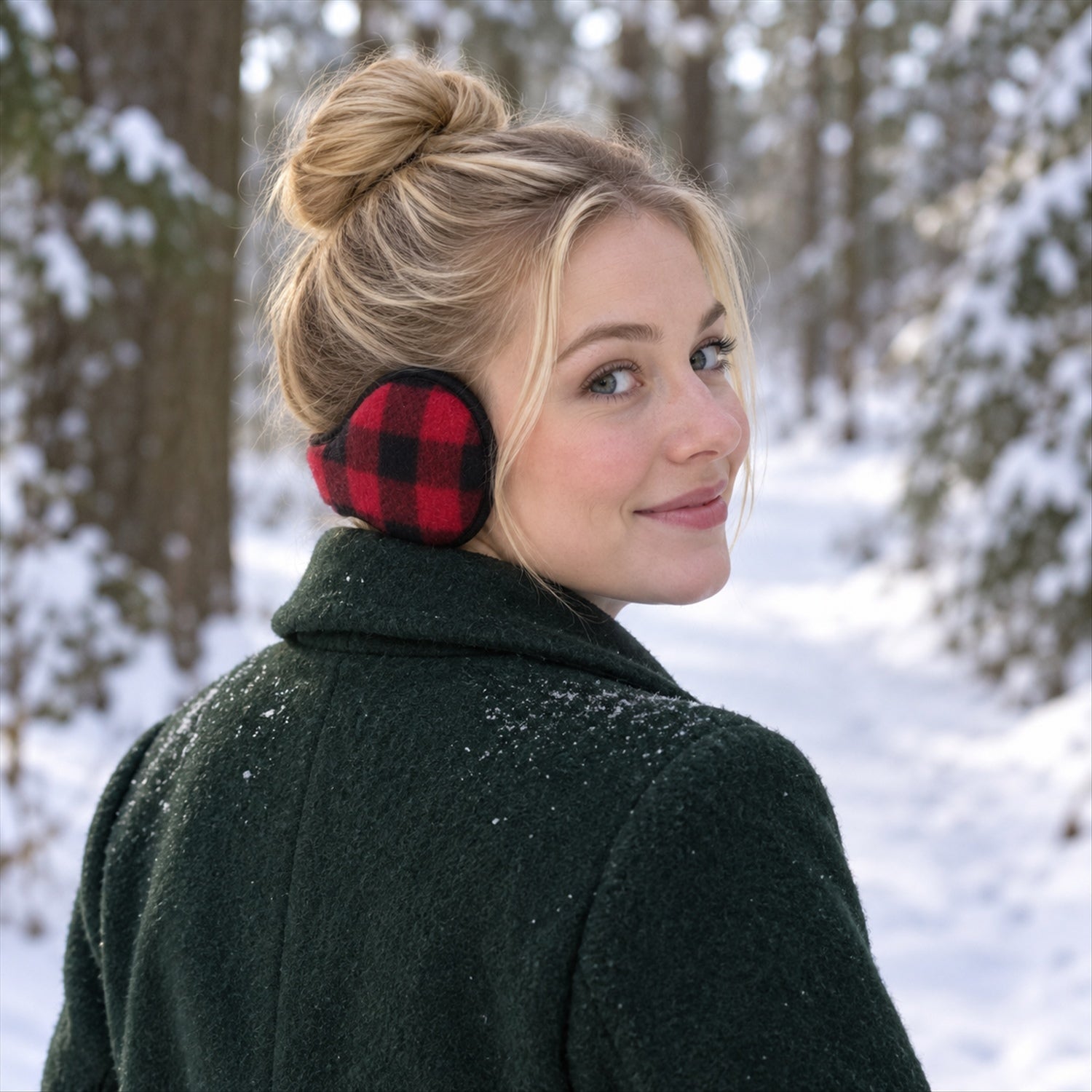 A woman with a bun wears red and black plaid ear warmers and a dark green coat in a snowy forest.