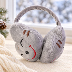 Grey plush earmuffs with a red embroidered smiley face on a cream knitted blanket, with a snowy window view in the