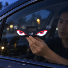 A person's hand holding the black evil eye light with glowing red and white eyes against a car window at night.
