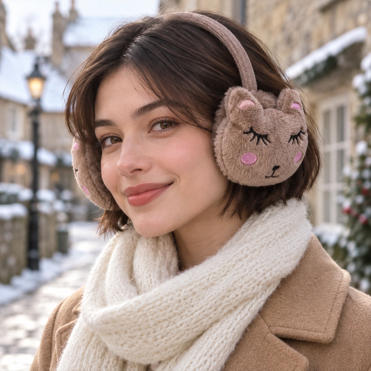 A smiling woman with short brown hair wears brown animal-face earmuffs and a cream scarf on a snowy street.