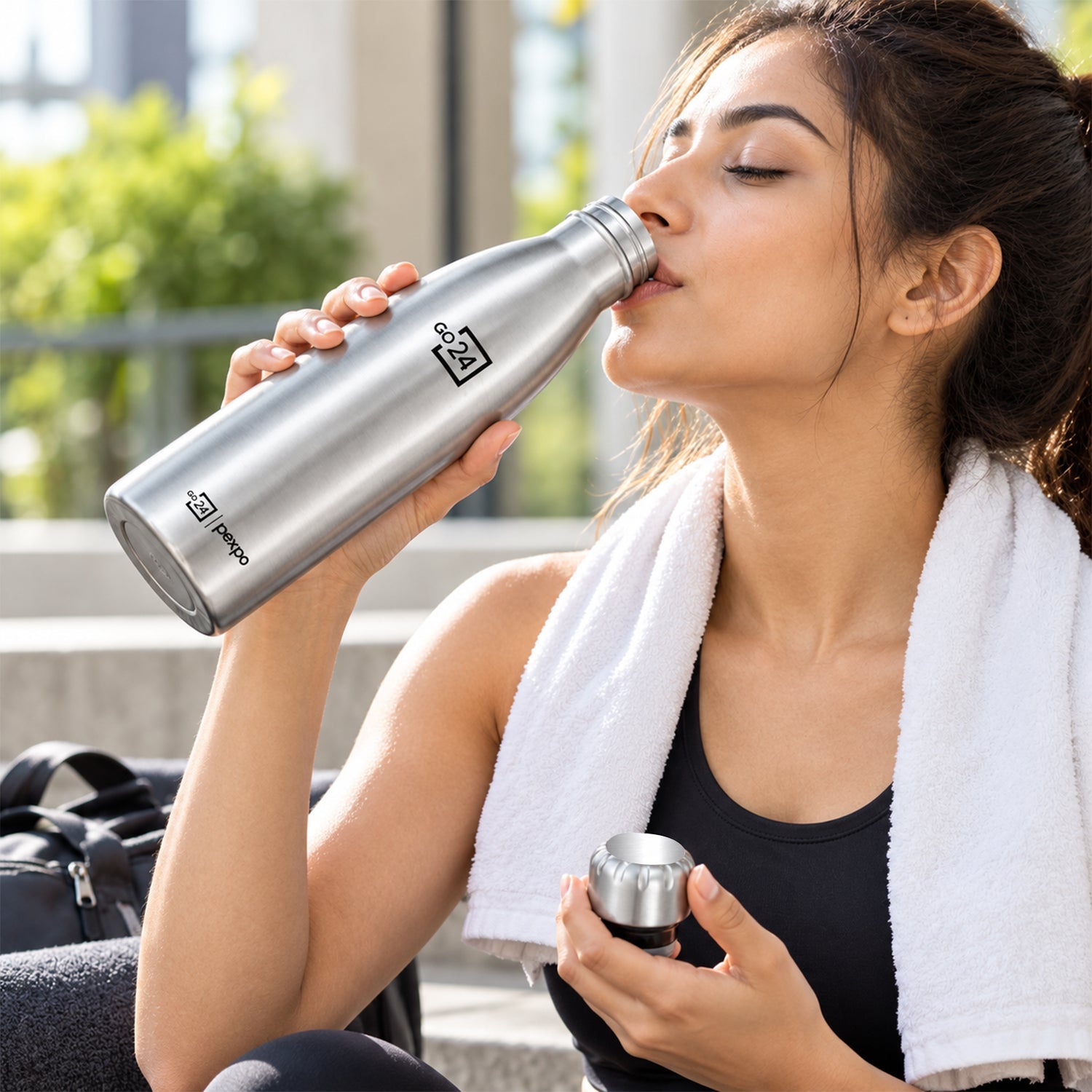 A woman in athletic wear drinks from a silver water bottle while sitting outdoors with a white towel.