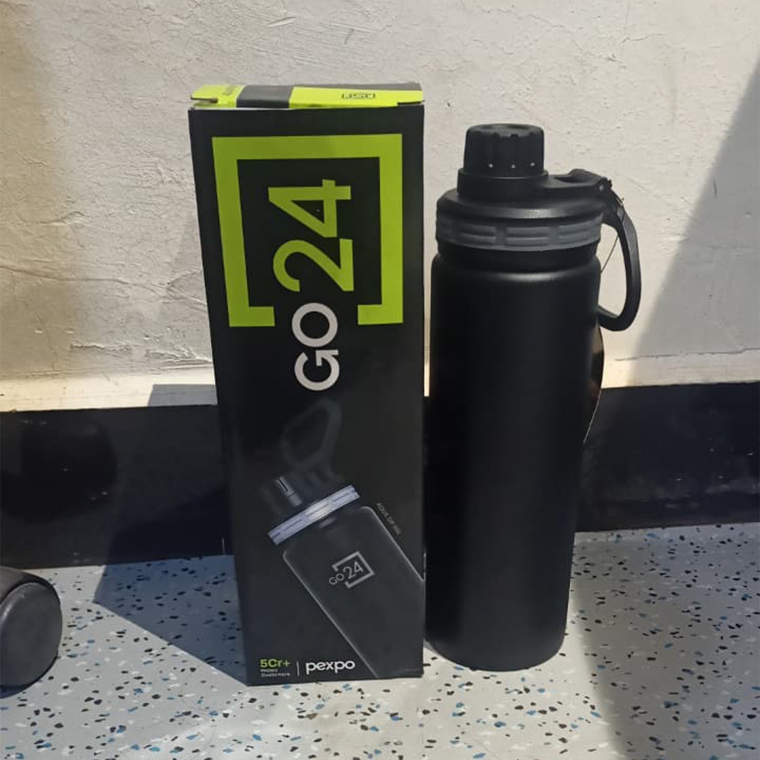 A black stainless steel bottle and its matching product box standing upright on a grey tiled floor.