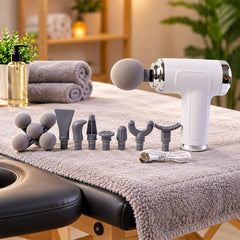 A white massage gun with various gray attachment heads, a USB cable, and rolled towels on a massage table.
