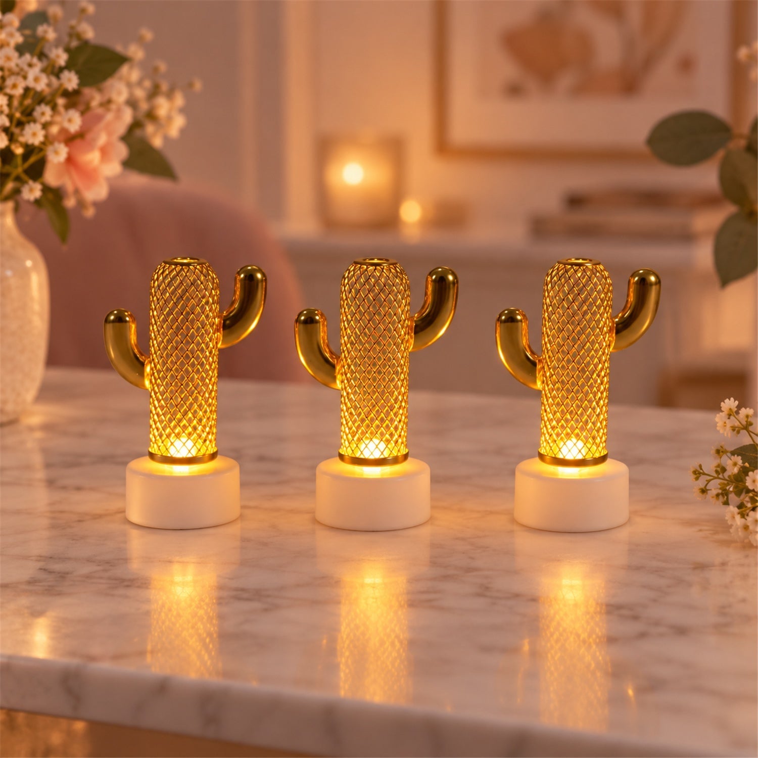 Three illuminated gold-tone cactus lights with white bases sit on a marble surface, surrounded by soft pink flowers.