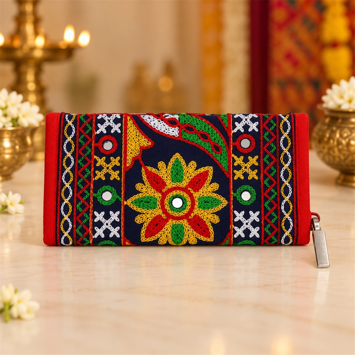 The embroidered clutch wallet rests on a light-colored surface, surrounded by small white flowers and blurred golden decor.